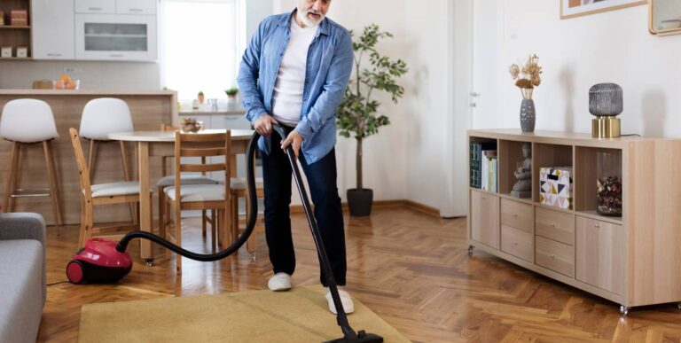 Na zdjęciu meżczyzna odkurzający dywan w pokoju. W tle stół oraz meble